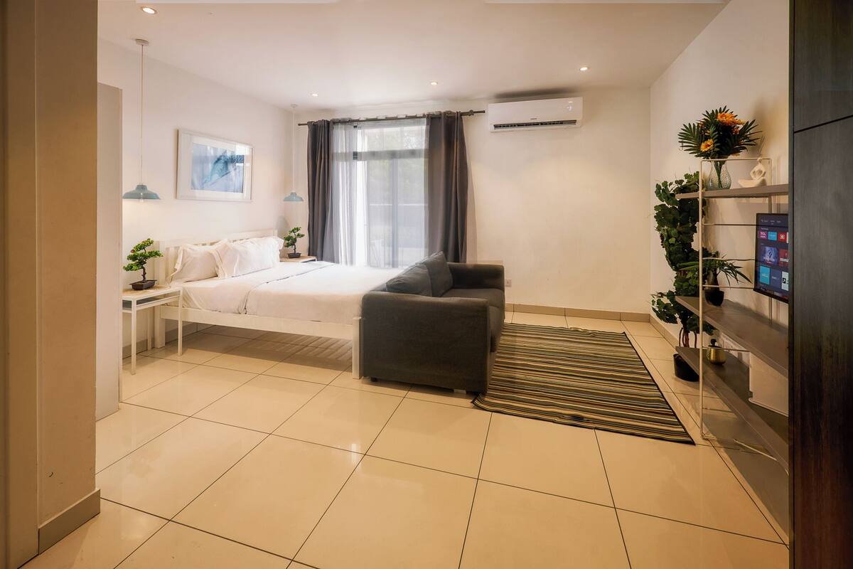 A modern hotel room with a bed, a sofa, a TV, a shelf with plants, and an air conditioning unit.