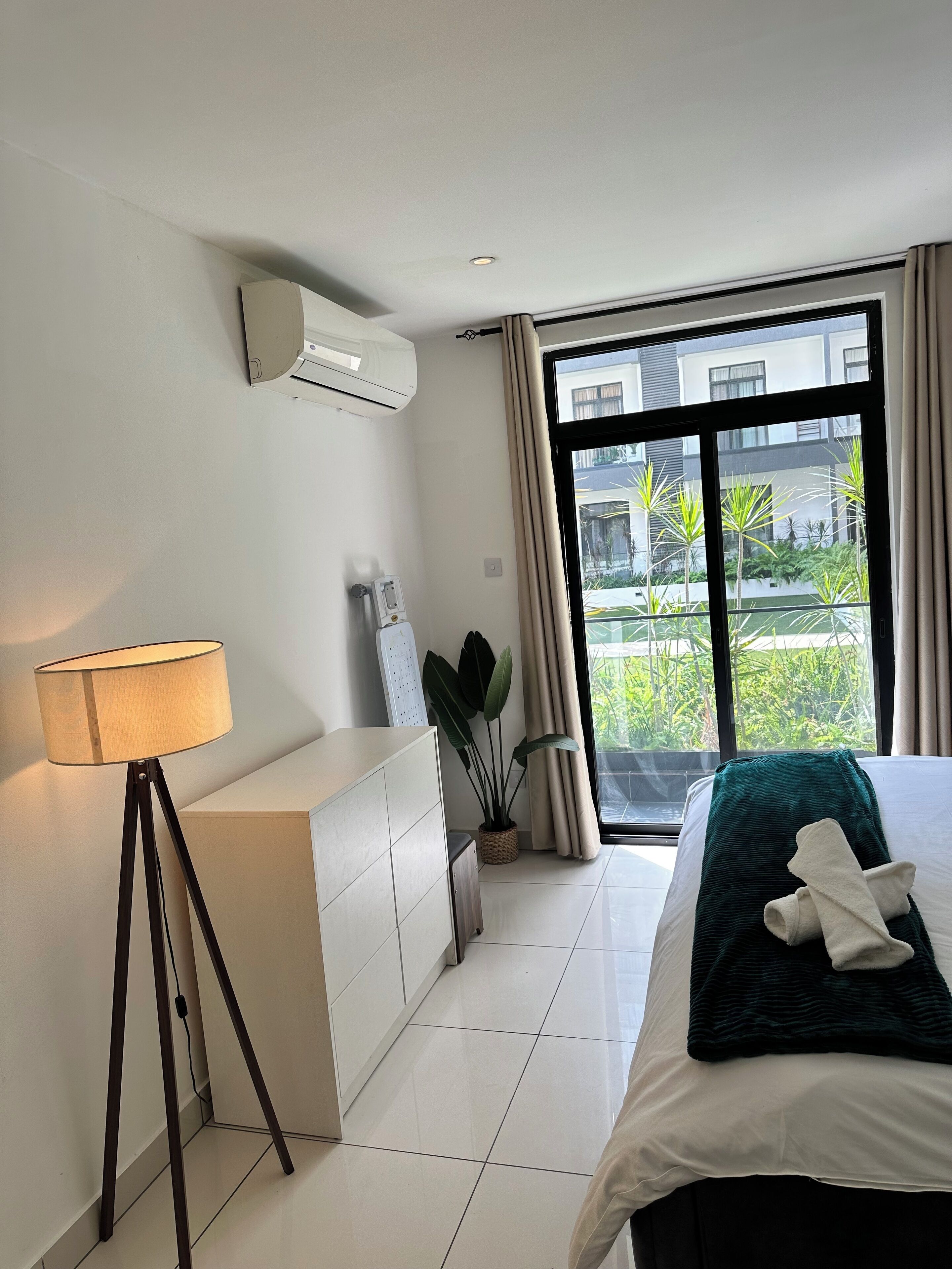 A hotel room with a bed, a bedside lamp, a potted plant, and a view of greenery outside.