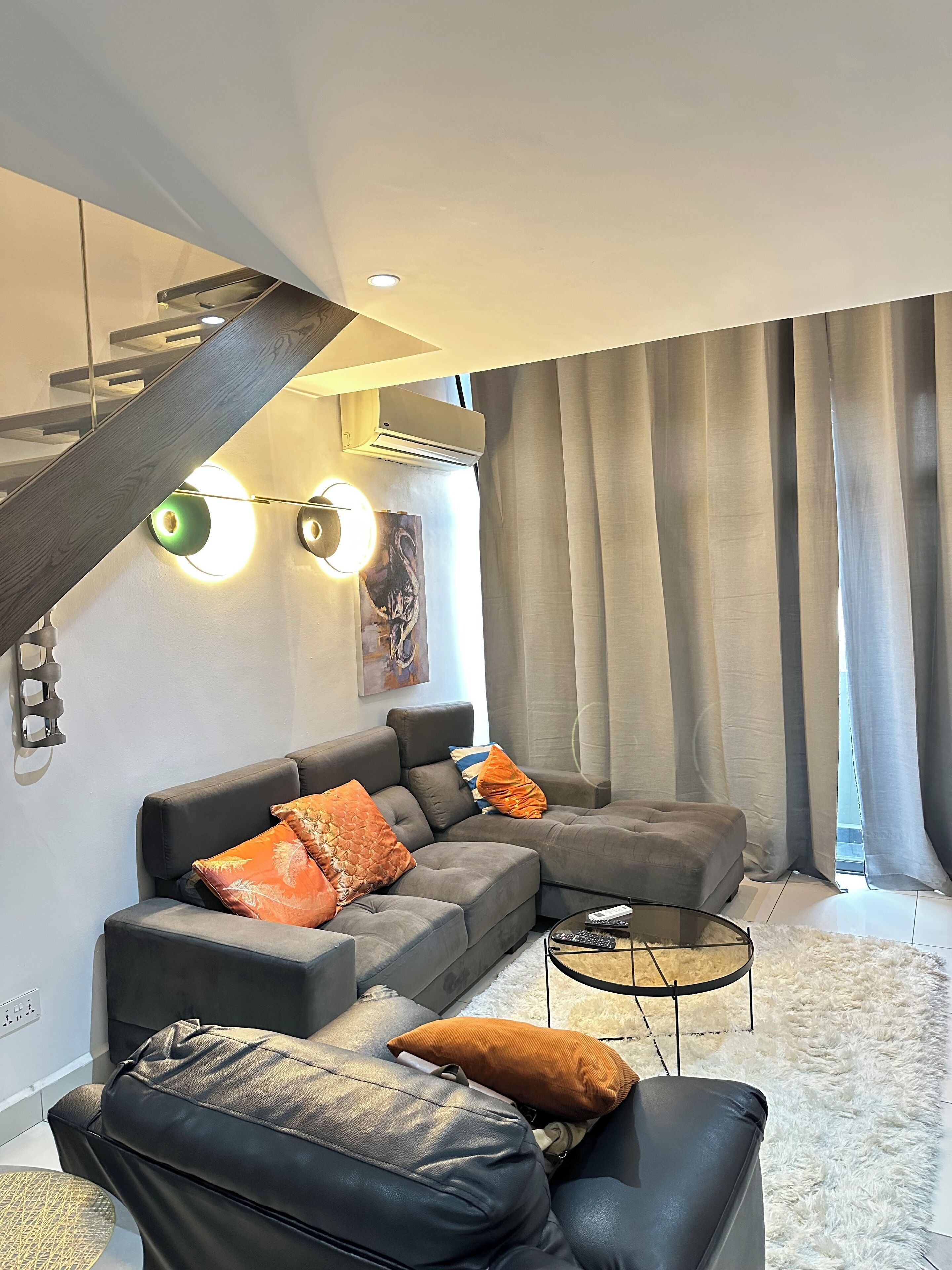 A modern living room with a grey sectional sofa, a black leather couch, and a glass coffee table. A staircase with a wooden railing is visible in the background.