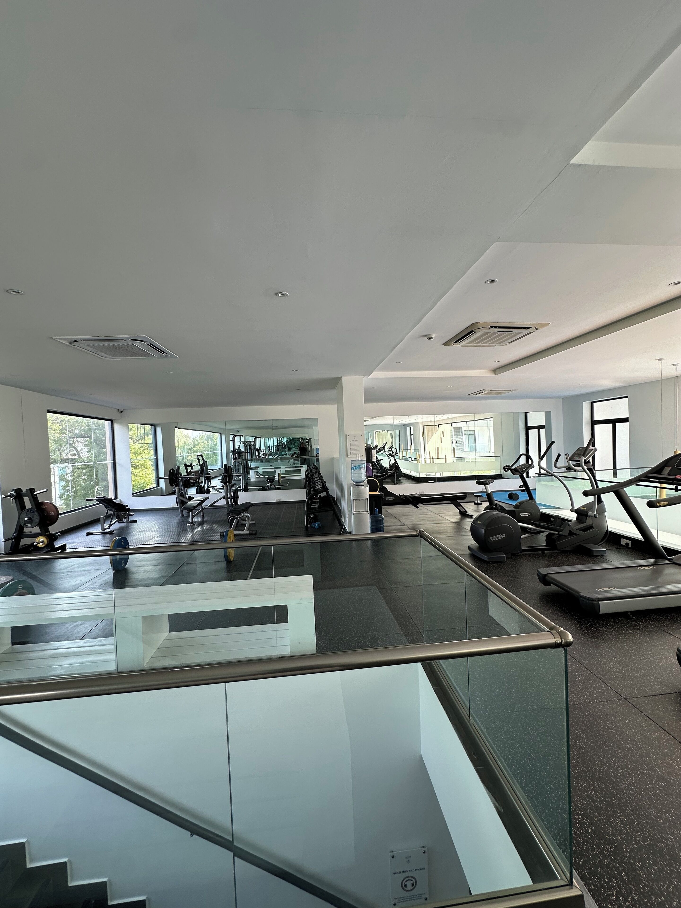 A modern gym with various exercise equipment, large windows, and a glass railing overlooking the space.