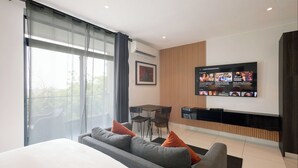 Habitación de hotel moderna con una cama grande, un sofá gris con cojines naranjas, una zona de comedor con mesa y sillas, y un televisor de pantalla plana montado en la pared que muestra una película.