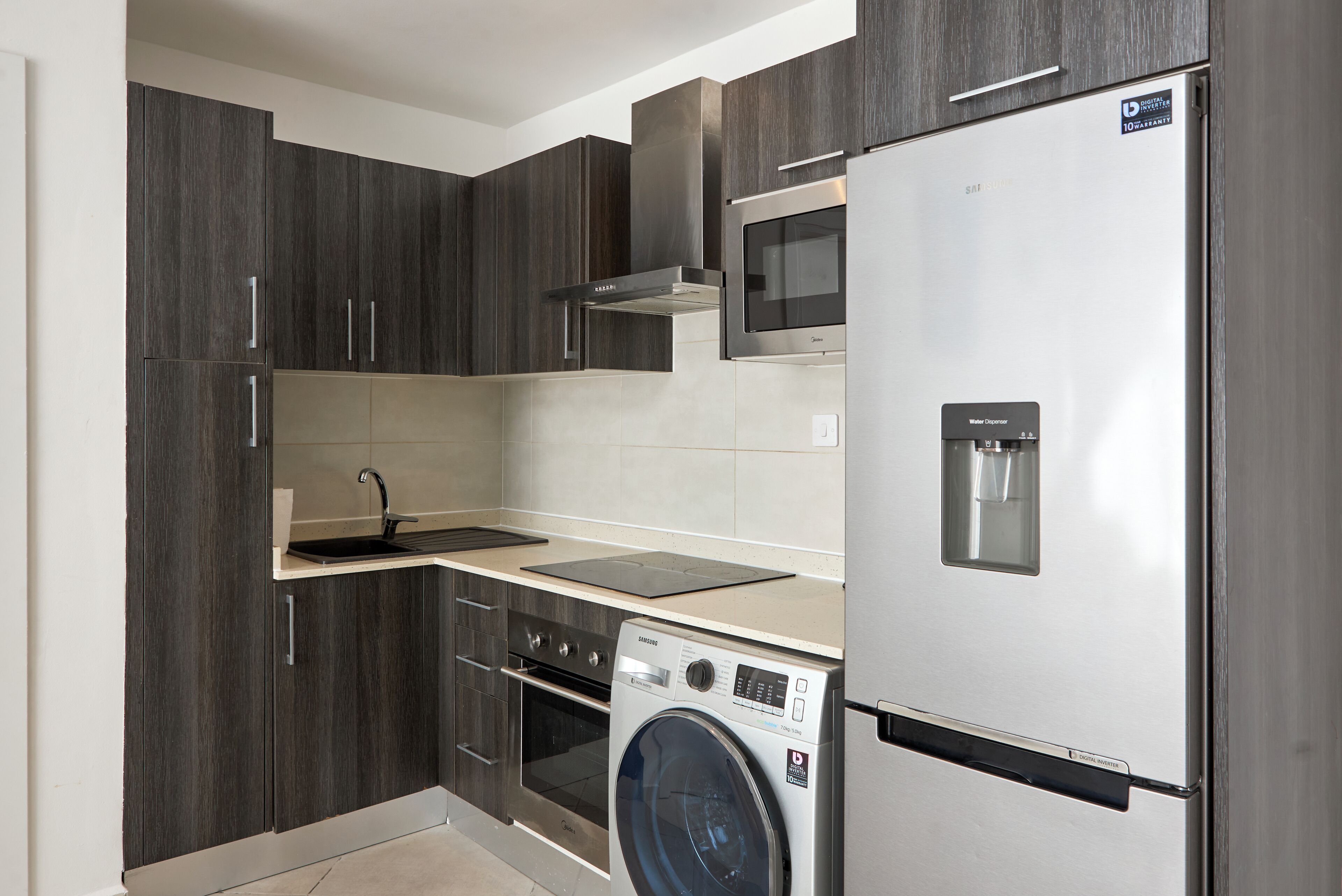 A compact kitchen with dark wood cabinets, a stainless steel refrigerator, a built-in oven, and a washing machine.