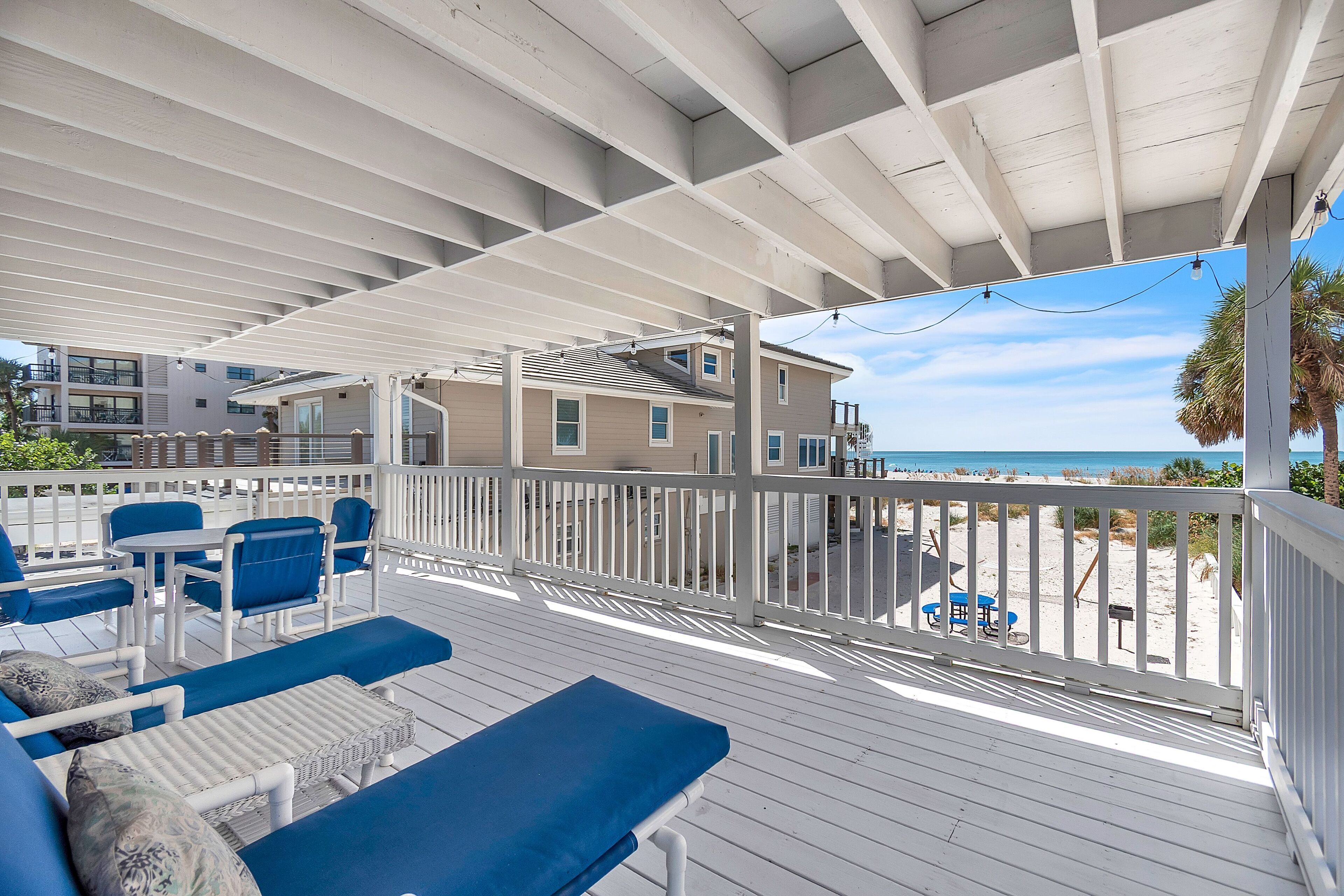 A covered outdoor deck with blue furniture, white railings, and a view of the beach.