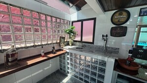 Un coin bar doté d’un comptoir en marbre, d’un évier et d’une bouteille de spiritueux. Une fenêtre à vitres teintées en rose et une plante en pot ornent le comptoir.