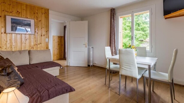 A room with a bed, a dining table with chairs, a radiator, and a window with curtains. - Hotel Sterne - Apartment with Balcony (Beatenberg)