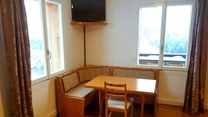 A room with a wooden table and chairs, a bench, a television mounted on the wall, and large windows with curtains. - Hotel Sterne - Apartment with Balcony (Beatenberg)