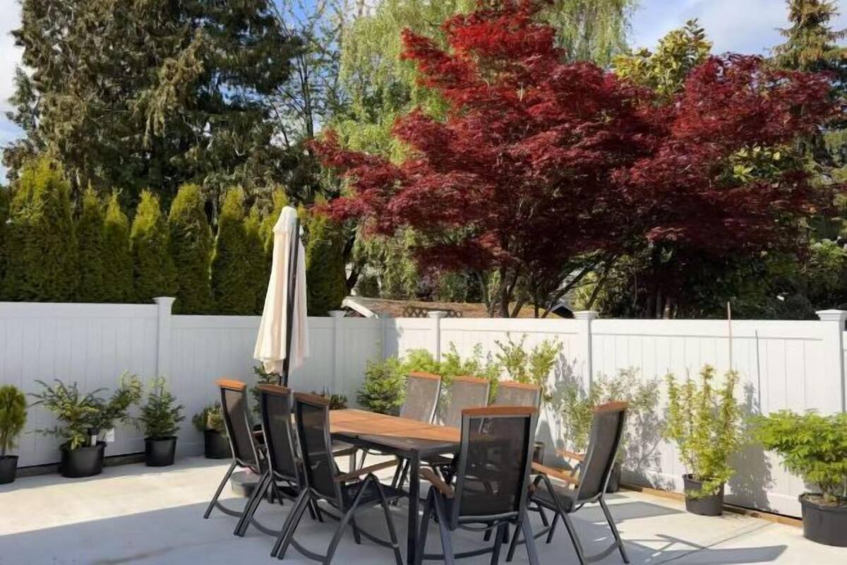Eine Terrasse mit Holztisch und -stühlen, umgeben von Kübelpflanzen und einem weißen Zaun.