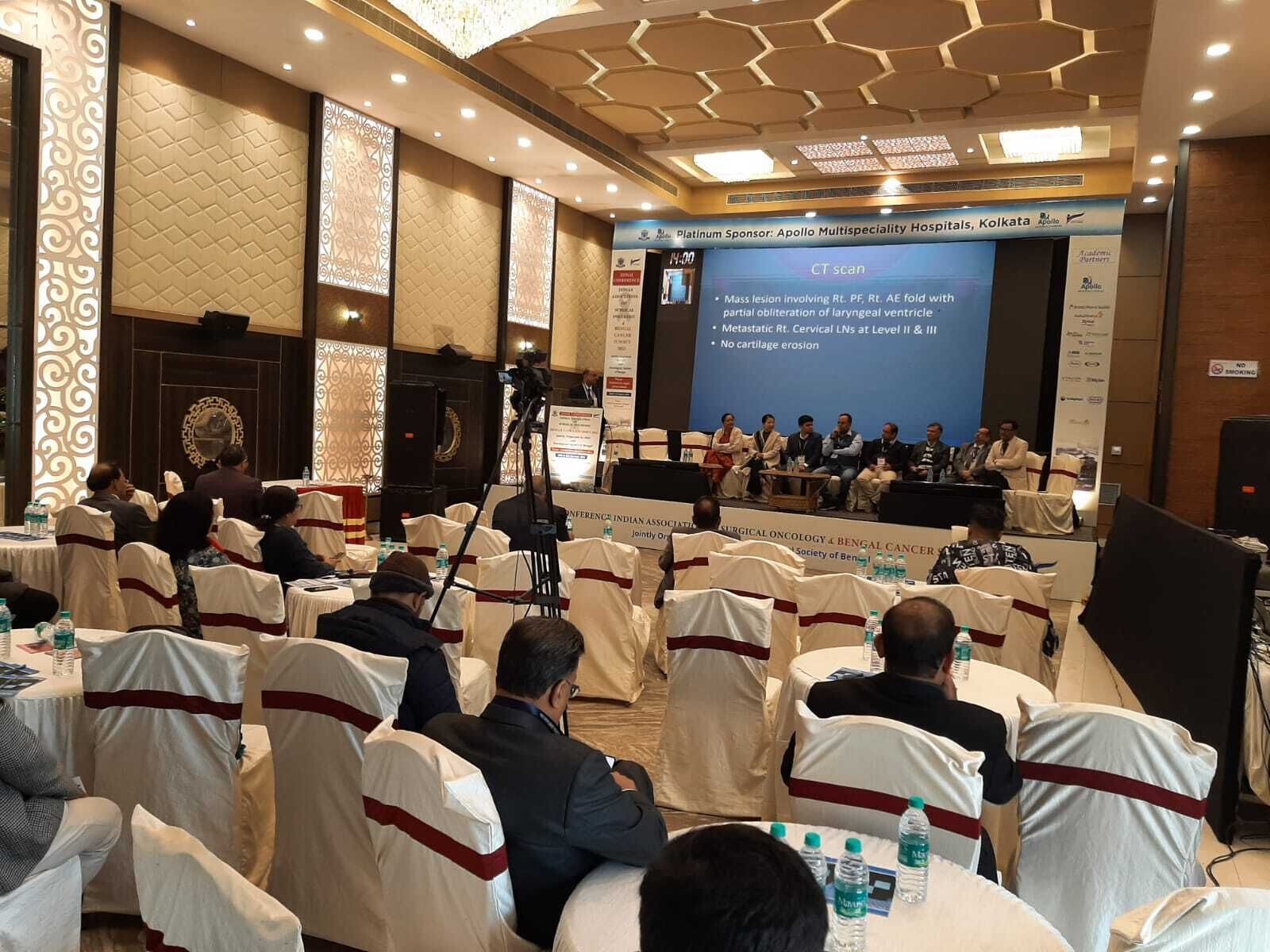 A conference room with attendees seated at round tables, a stage with a presentation screen, and a chandelier.