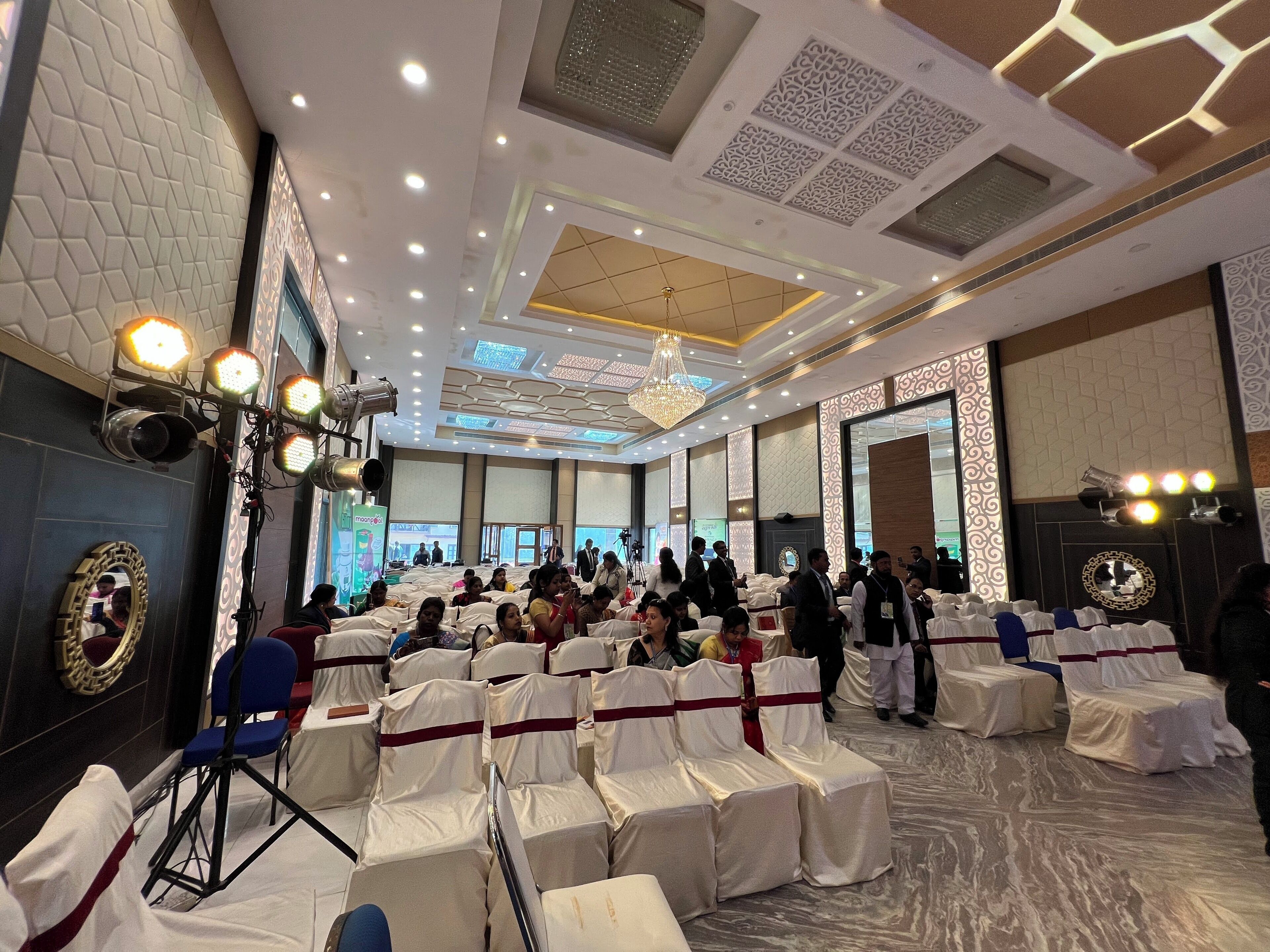 A banquet hall with rows of white-covered chairs, a chandelier, and a patterned ceiling.