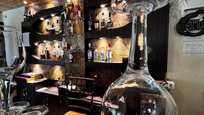 Zona de bar con estanterías llenas de botellas de licor, una botella grande de vino y varios vasos sobre una mesa.