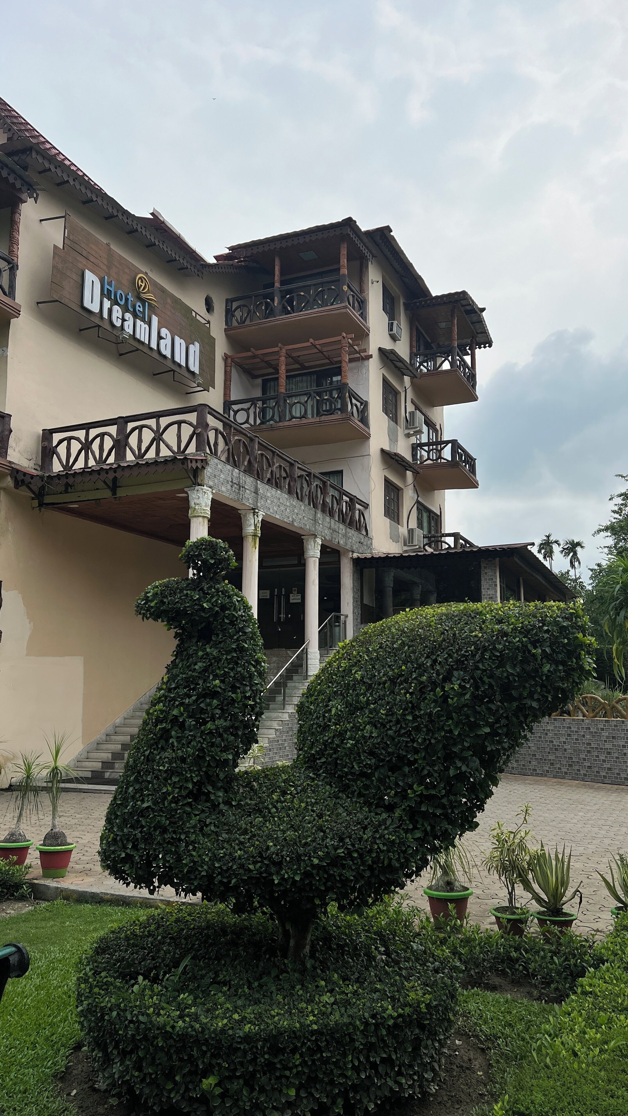 A multi-story building with a sign reading "Hotel Dreamland." It features balconies with intricate railings, a staircase leading to the entrance, and a well-maintained garden with various plants.