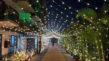 Un camino bordeado por plantas en macetas y luces colgantes, que conduce a la entrada de un edificio.