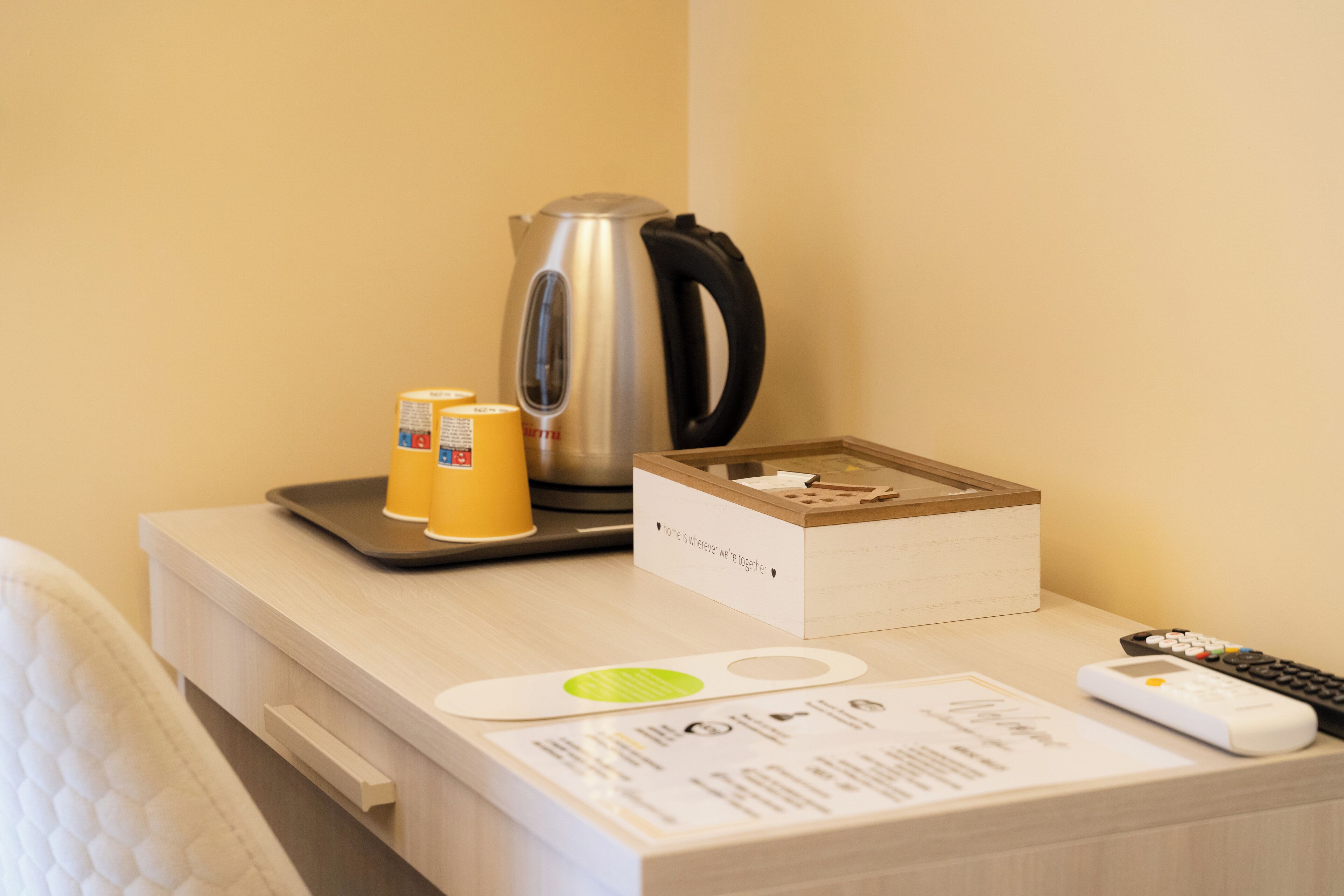 Een hotelkamer met een houten nachtkastje, een waterkoker, een dienblad met twee kopjes, een doos met een briefje, een afstandsbediening en een menukaart.