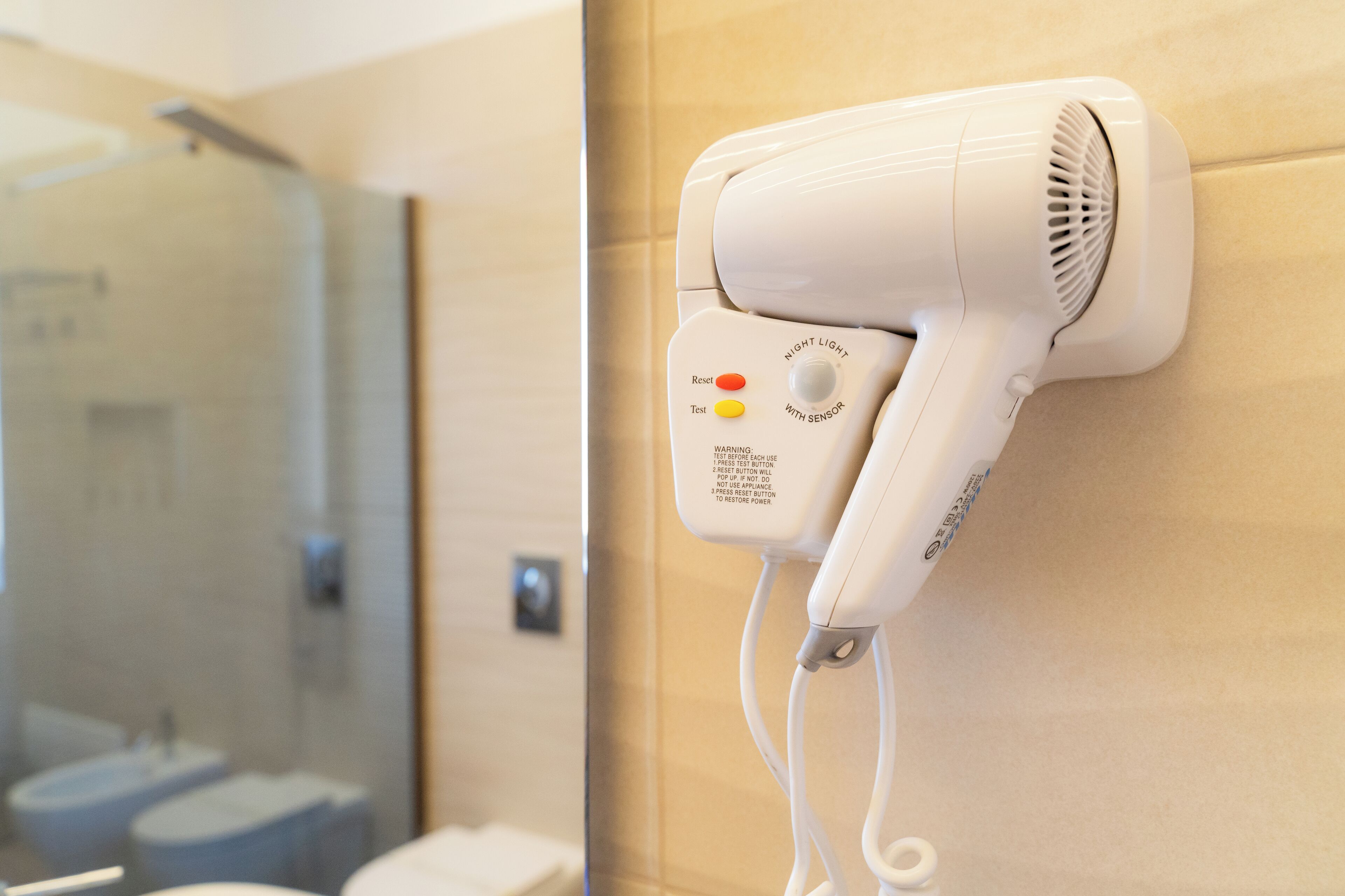 Un sèche-cheveux fixé au mur dans une salle de bain, avec une douche en verre en arrière-plan.