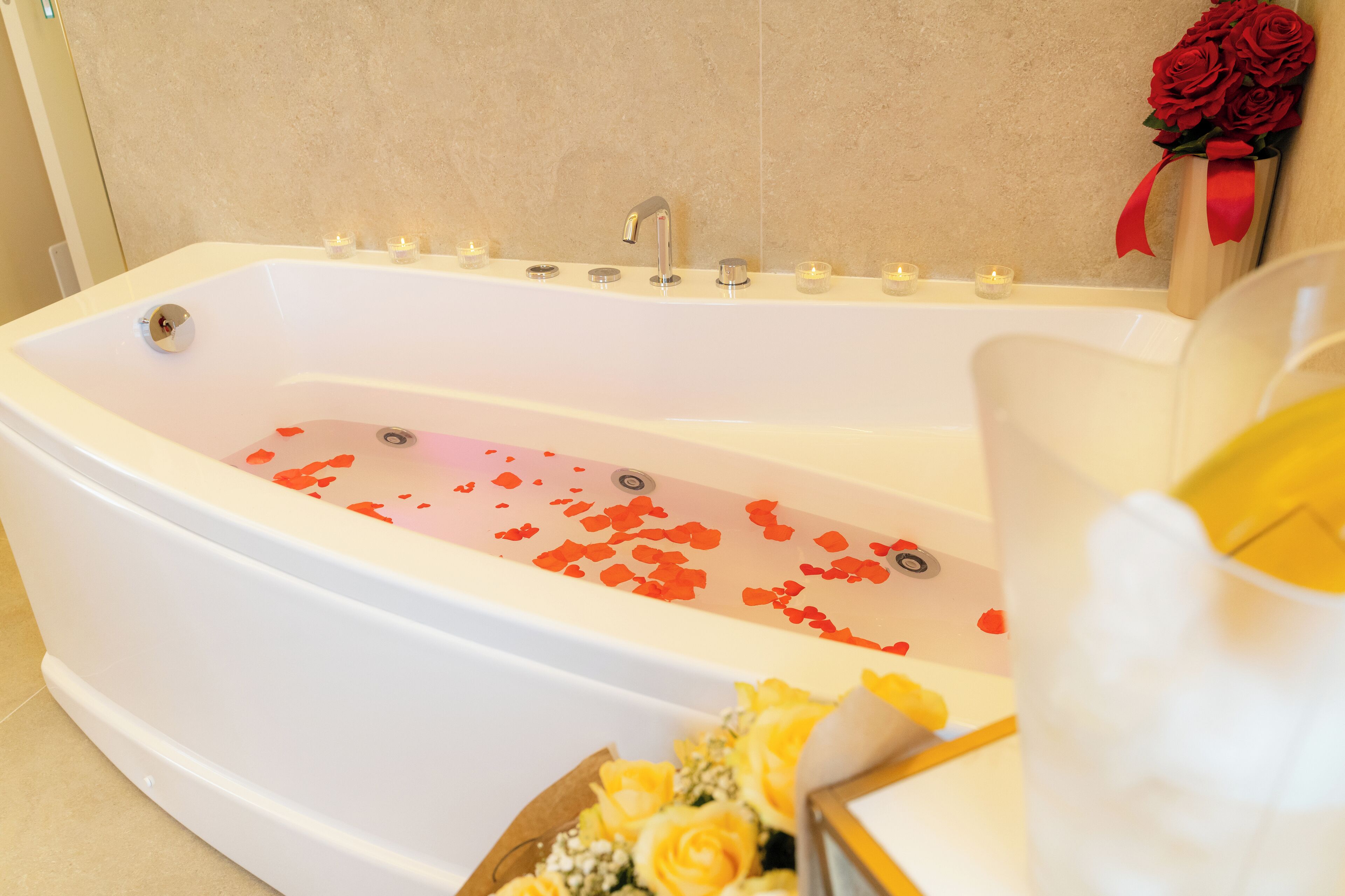 Eine Badewanne mit Rosenblättern und Kerzen, ein Strauß gelber Rosen und eine Vase mit roten Rosen.