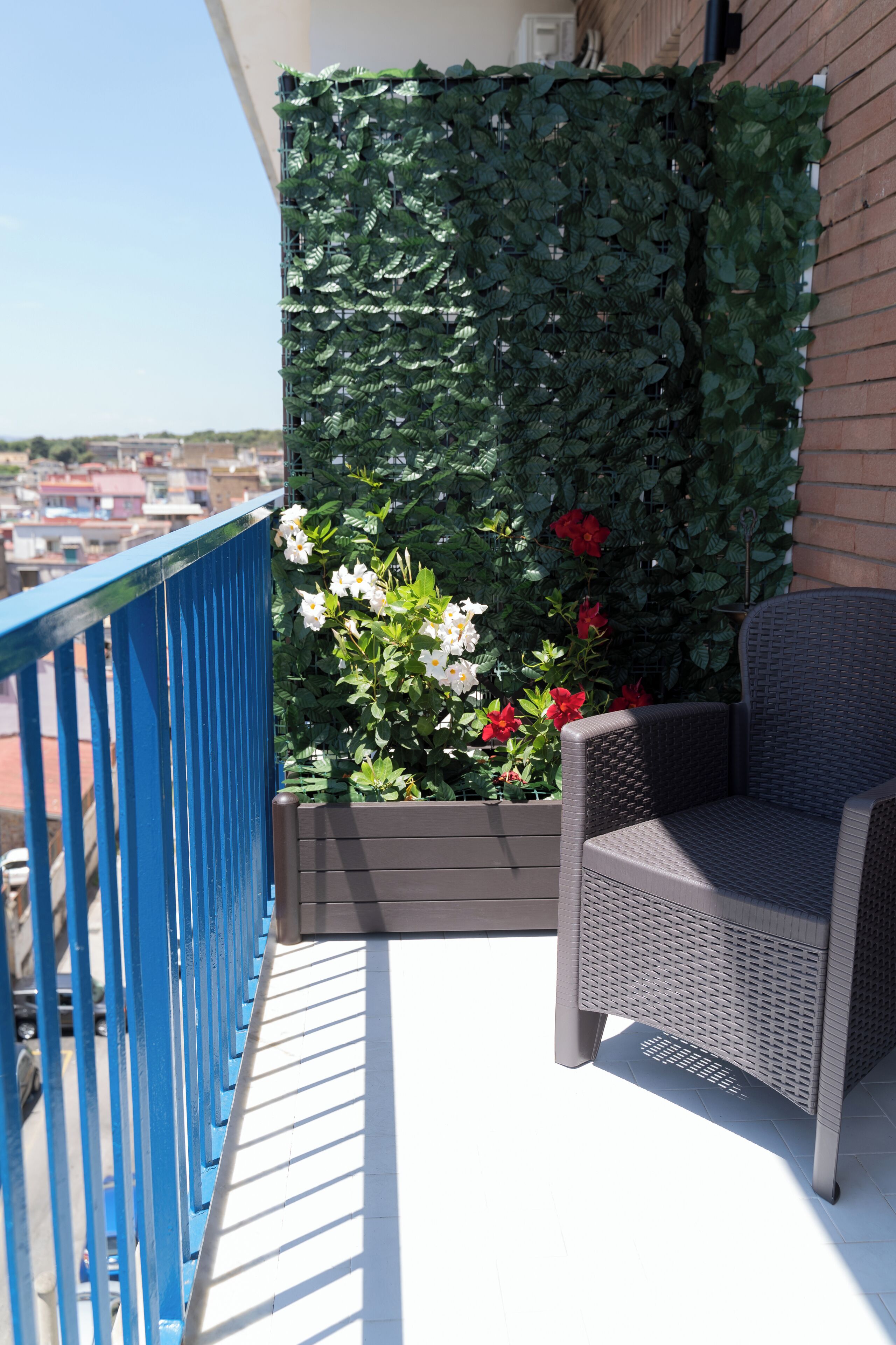 Un balcón con baranda azul, una silla de mimbre y una maceta con flores.