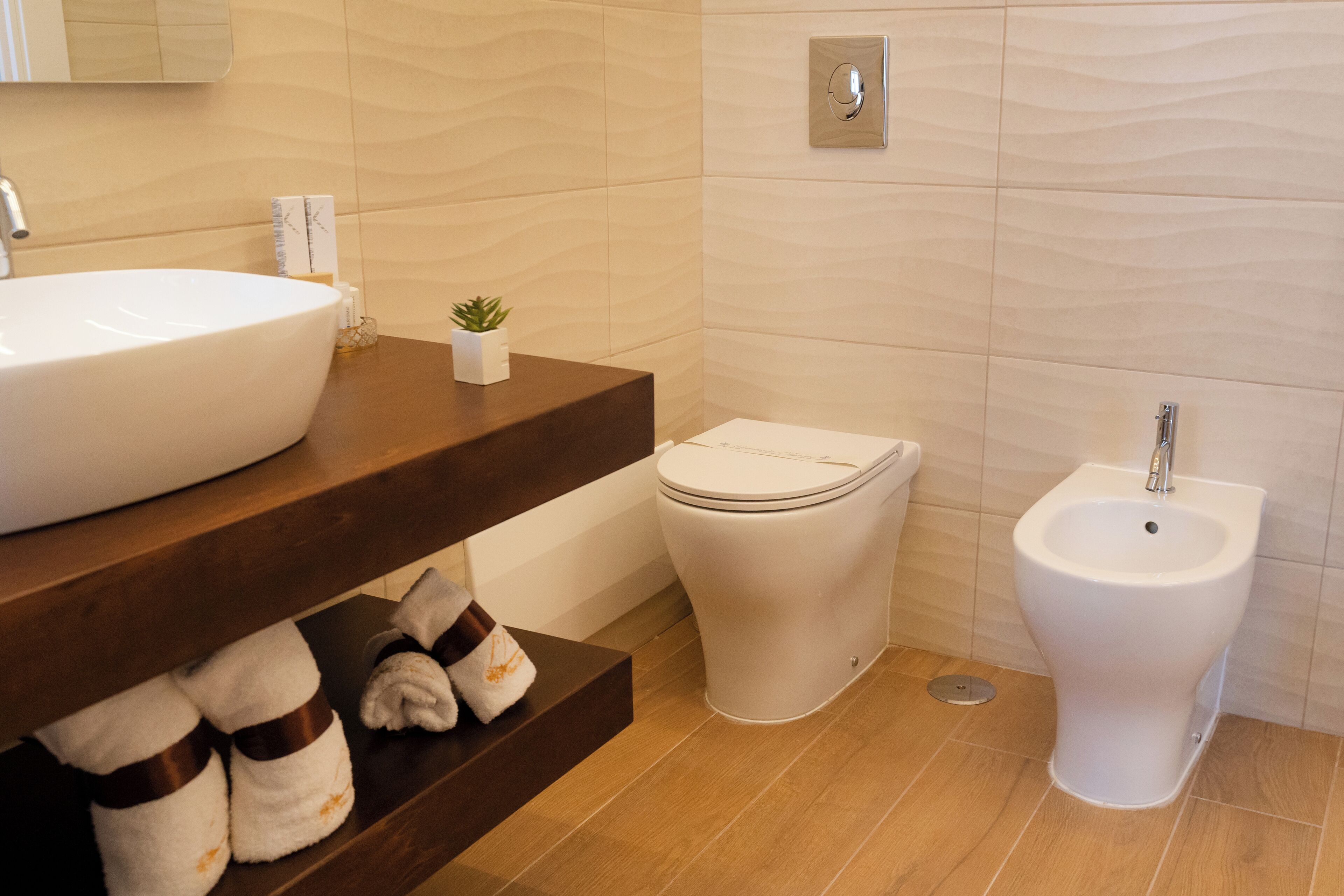 A modern bathroom with a large white sink, a toilet, and a bidet. There are towels on a shelf and a small plant on the counter.