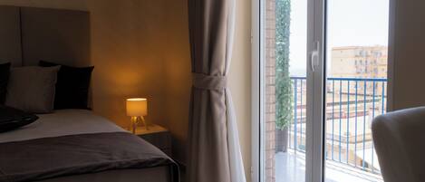 A hotel room with a bed, bedside table, lamp, curtains, and a view of the outside balcony.