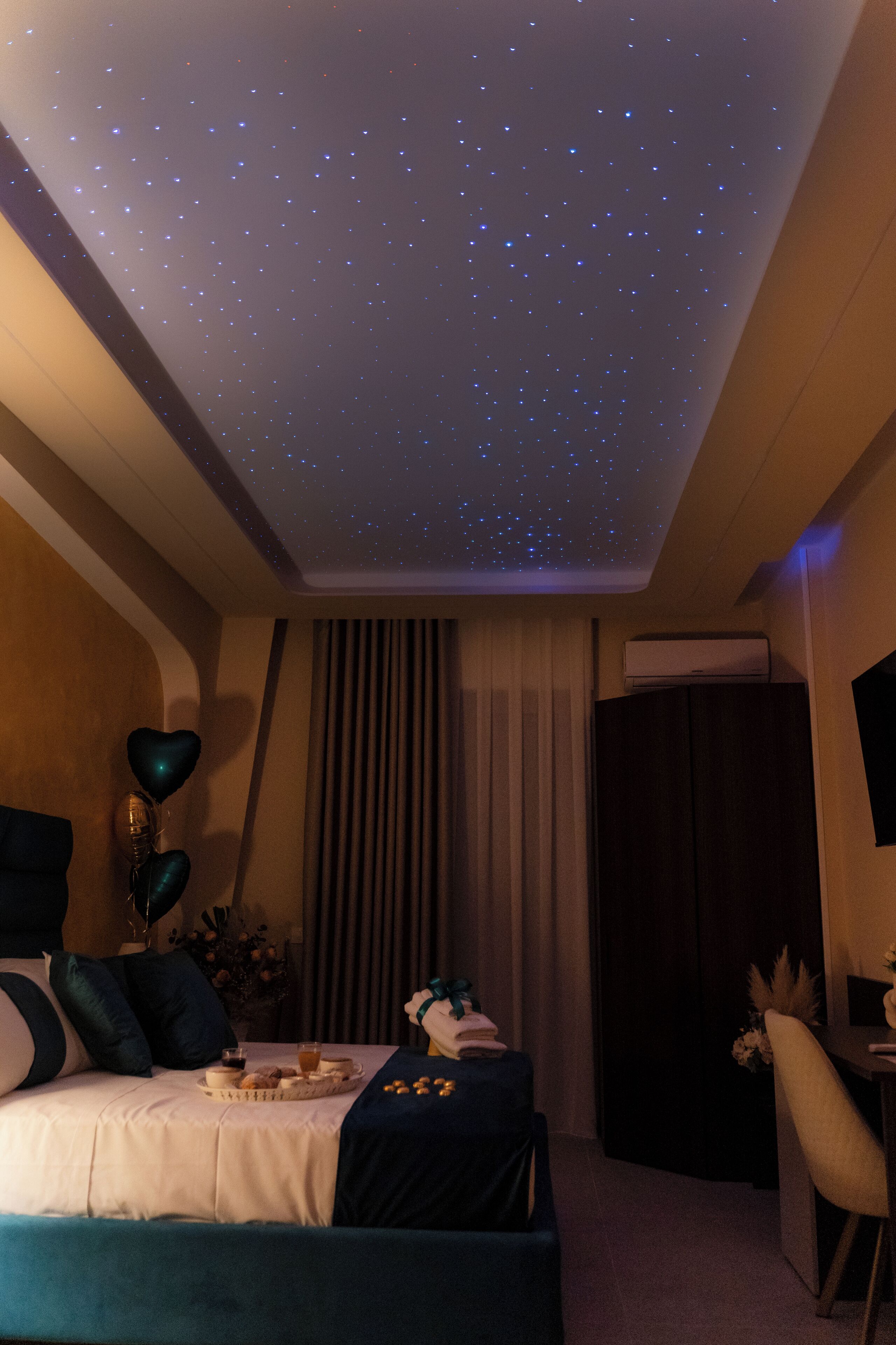 A hotel room with a starry ceiling, a bed with a blue headboard, a bedside table with a lamp, and a window with curtains.