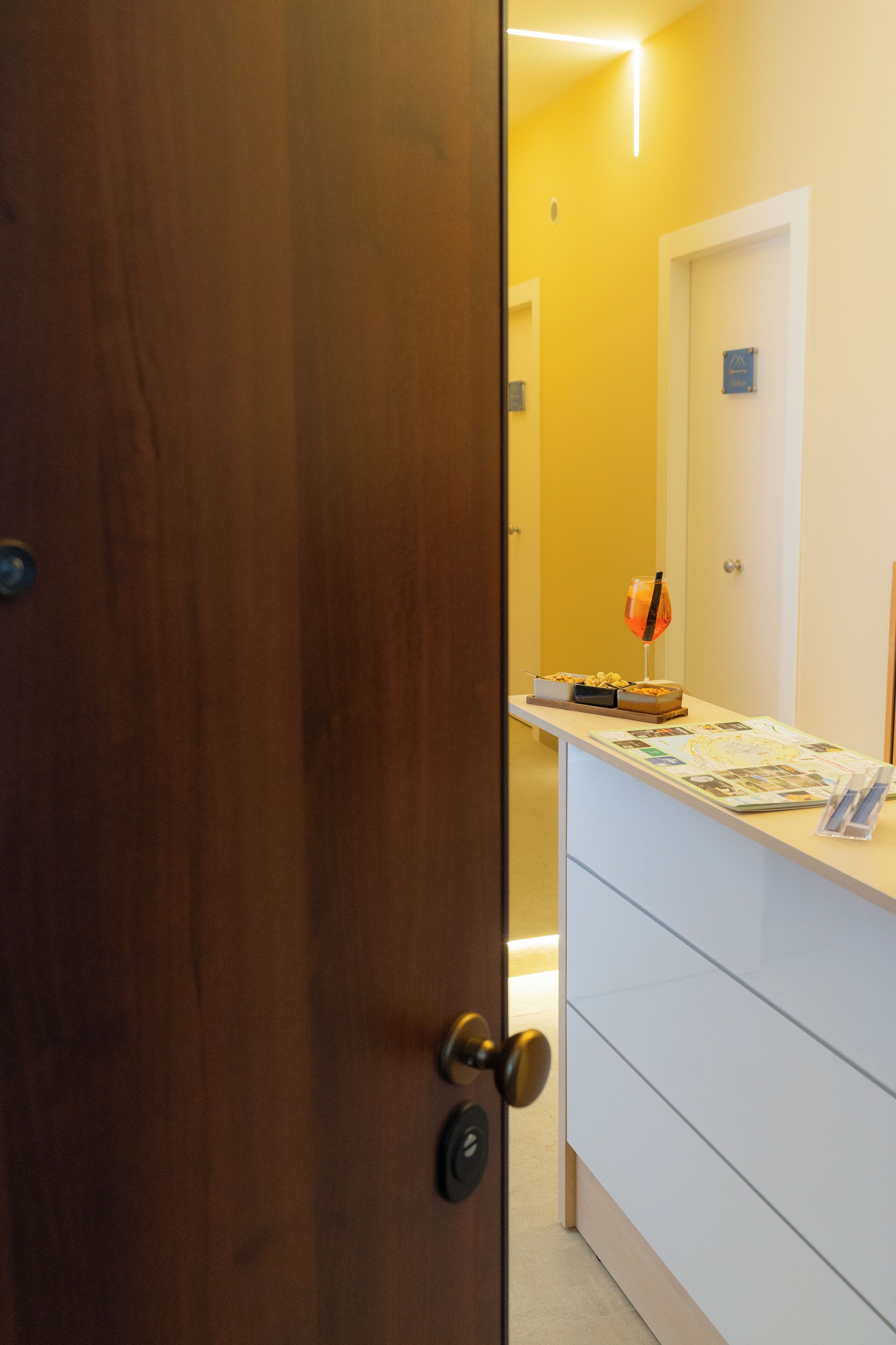 A hotel room with a wooden door, a white dresser, a small table with a glass, and a map.