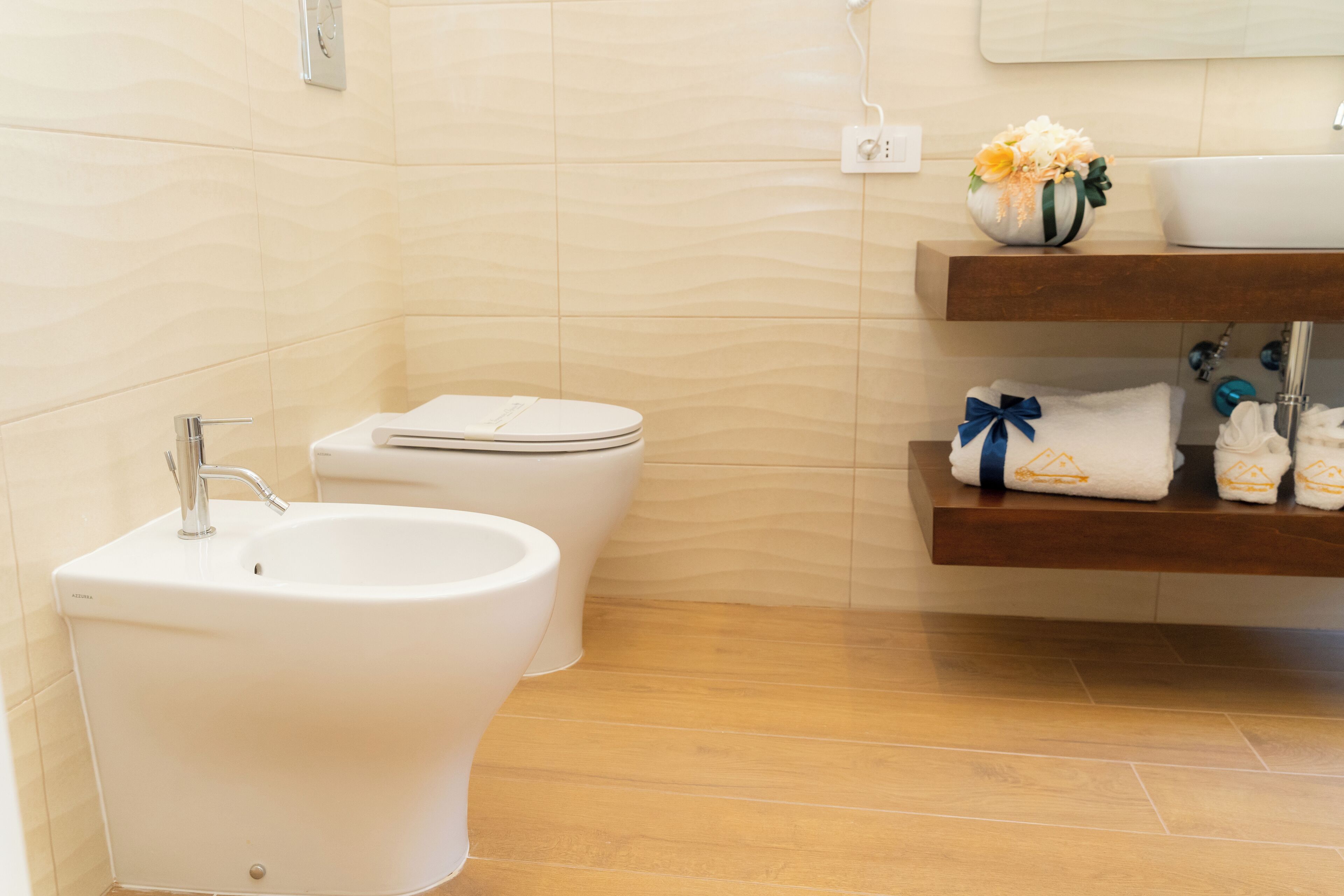 A bathroom with a bidet, toilet, and a shelf with towels and a flower vase.