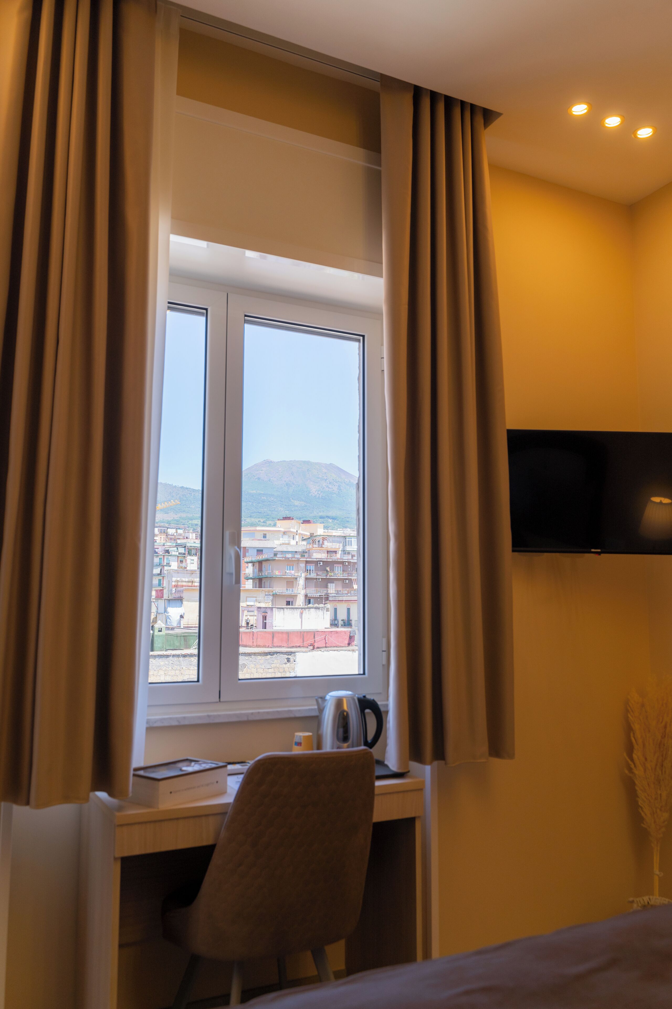 Une chambre d’hôtel équipée d’un bureau, d’une chaise et d’un lit. On y trouve une fenêtre avec des rideaux, un téléviseur à écran plat fixé au mur, et une vue sur les bâtiments et les montagnes environnants.