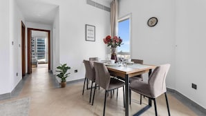 A dining area with a wooden table set for four, surrounded by chairs, a potted plant, and a window with curtains. - StayShort - Stylish Barsha Heights Retreat with Swimming Pool for 2 (Barsha Heights)