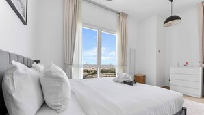 A modern bedroom with a large bed, a dresser, and a view of the city through the window. - StayShort - Stylish Barsha Heights Retreat with Swimming Pool for 2 (Barsha Heights)