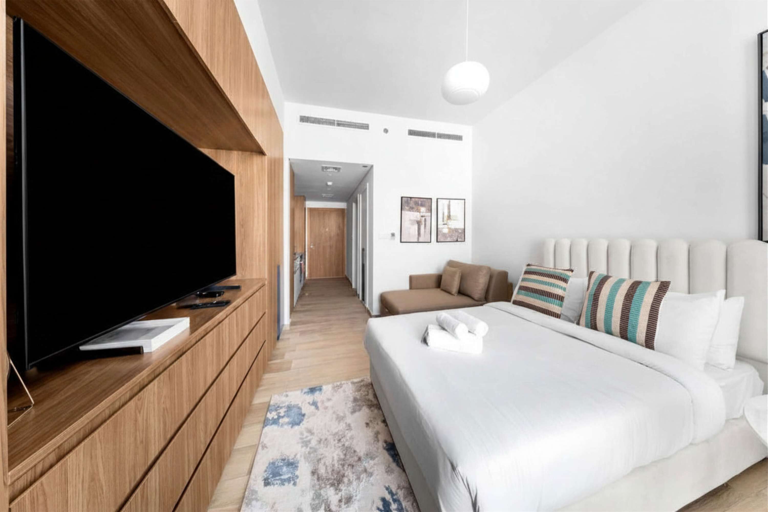 Ett modernt hotellrum med en platt-TV, en stor säng med många kuddar, en träkonsol och en hall som leder till ett annat rum.