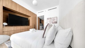 Une chambre d’hôtel moderne avec un grand lit, un téléviseur à écran plat fixé sur un panneau en bois, une petite table ronde et une vue sur la cuisine.