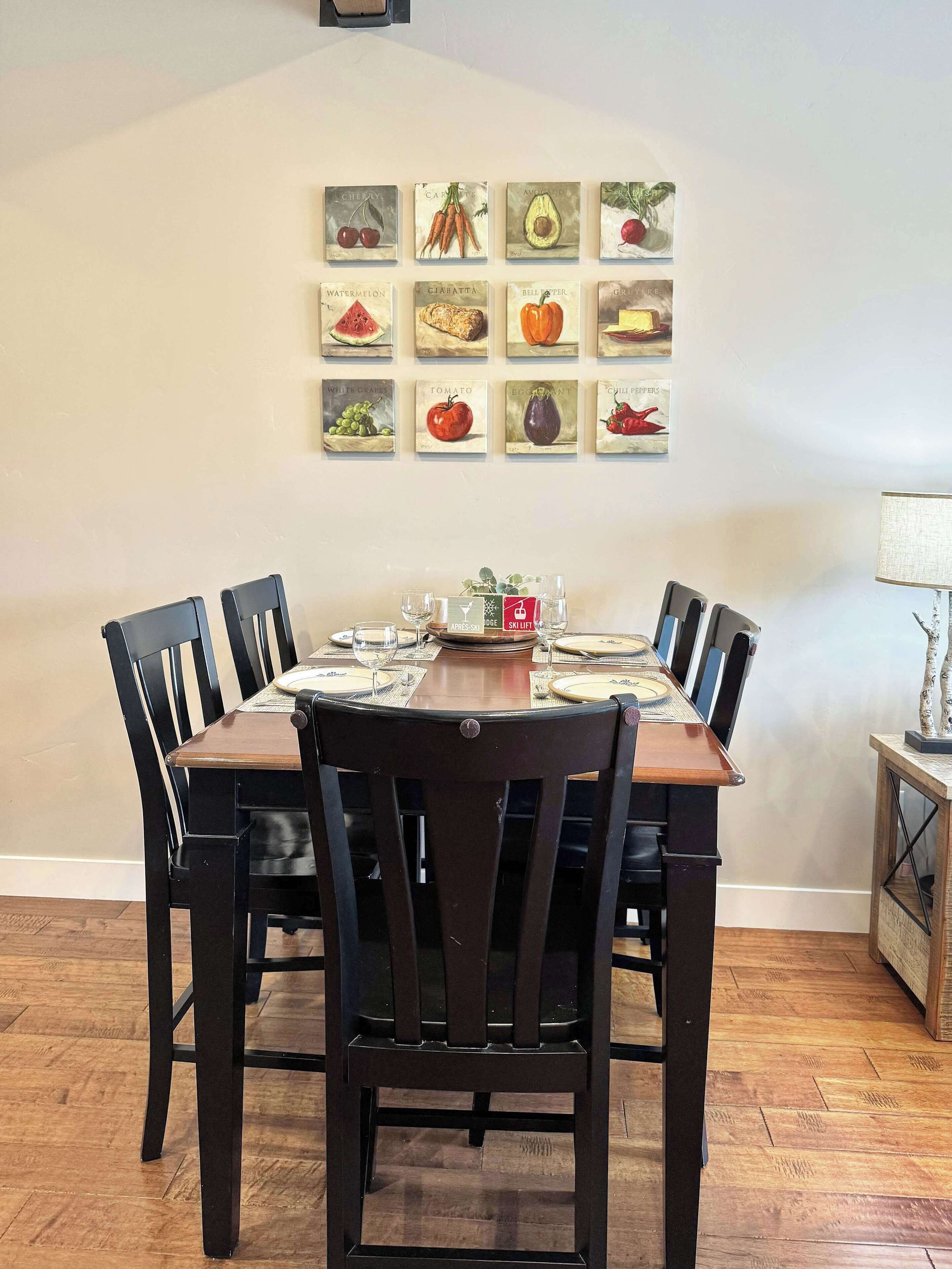 Un comedor con una mesa de madera, sillas negras y una obra de arte con motivos de frutas colgada en la pared.