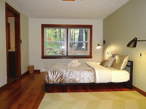 A bedroom with a bed, a window with a forest view, a bedside lamp, and a door.
