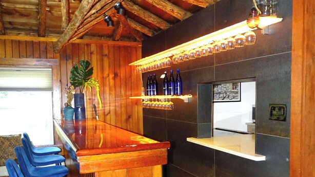 A wooden bar area with stools, a shelf with bottles, and a hanging light fixture.
