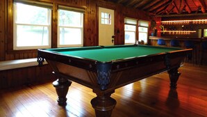A pool table in a room with wooden walls and a sloped ceiling. There are windows with blinds, a bar area with lights, and a bench along the wall.