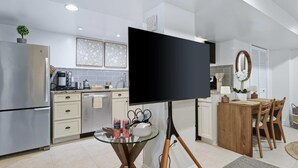 Cuina moderna amb un gran televisor de pantalla plana sobre un suport, zona de menjador amb taula i cadires de fusta, i una taula auxiliar petita amb una planta i parell de llaunes.