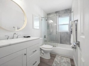 A modern bathroom with a white vanity, a round mirror, a toilet, and a glass-enclosed shower area. - Waterfront-Family & Pet Friendly for 6 (Apollo Beach)