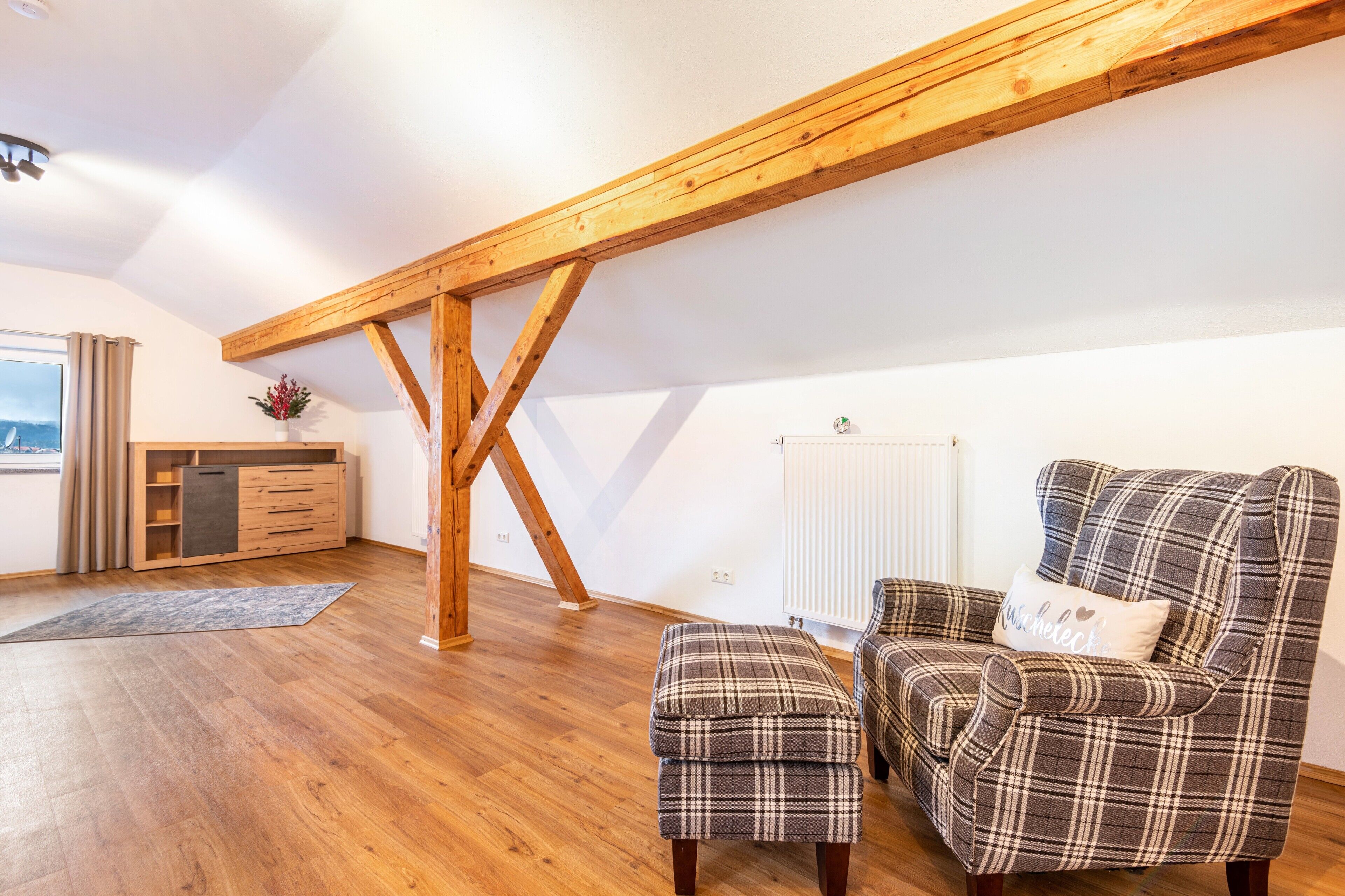 Een loft-achtige kamer met houten balken, een bank, een voetenbankje en een kleine tafel met een bloevas.