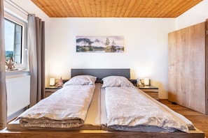 Un lit double avec une tête de lit, deux tables de chevet avec des lampes, une fenêtre avec des rideaux et un tableau représentant un paysage accroché au mur.