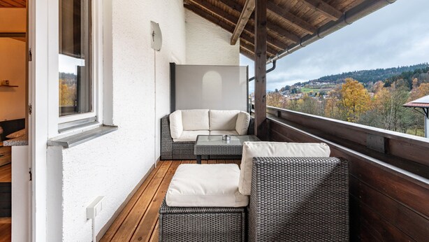 En balkon med fletningsmøbler, en hvid væg og udsigt over et kuperet landskab.