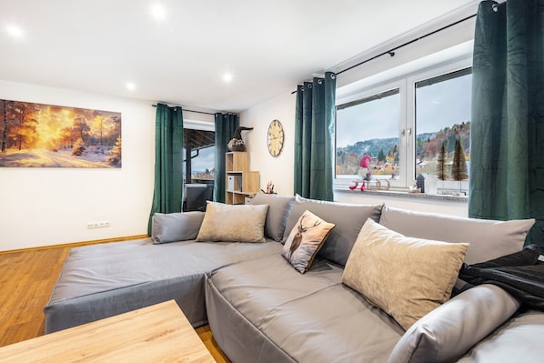 Un salon moderne avec un canapé gris, une table basse en bois, un tableau et une grande fenêtre avec des rideaux.