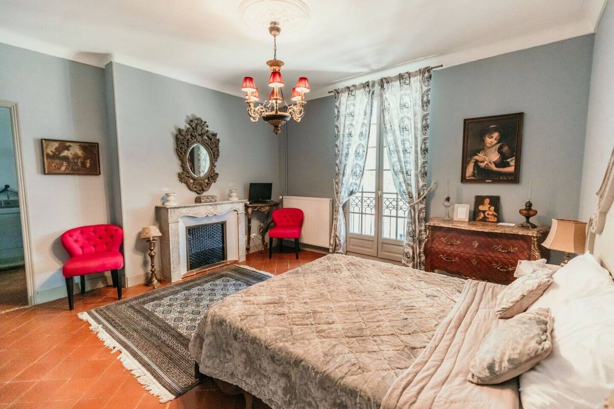 A bedroom with a large bed, a red chair, a fireplace, and a chandelier.