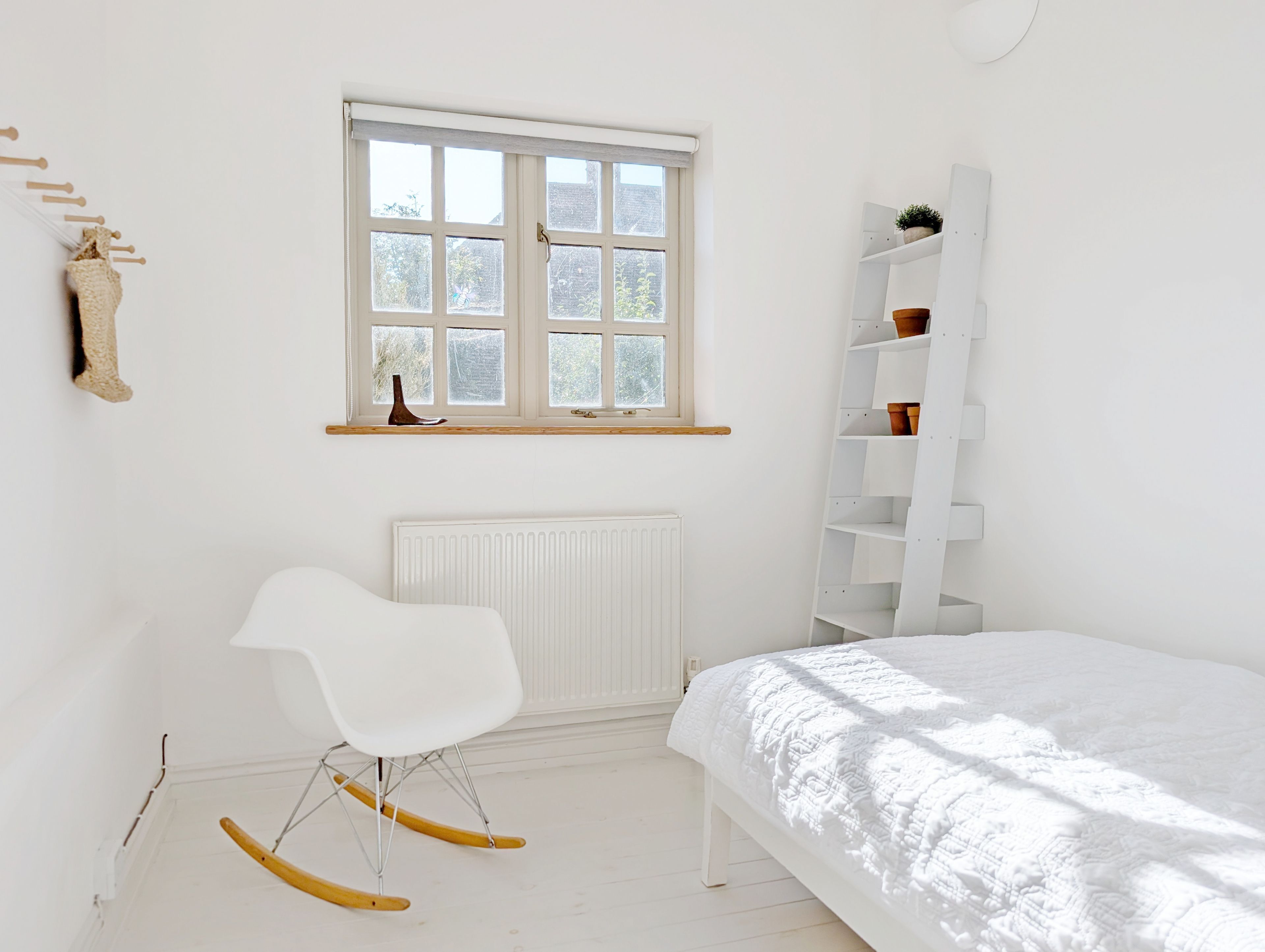 Un dormitorio con una cama blanca, una silla blanca, un radiador, una ventana y un estante blanco con plantas en macetas.