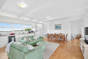 A spacious living area with a green sofa, dining table, and kitchen area with white cabinets and a view of the outdoors.