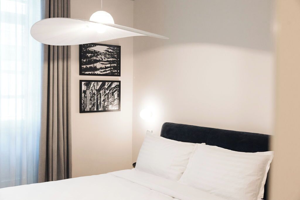 Hotellihuone, jossa on sänky, suuri valkoinen riippuvalaisin ja kaksi mustavalkoista valokuvaa seinällä.