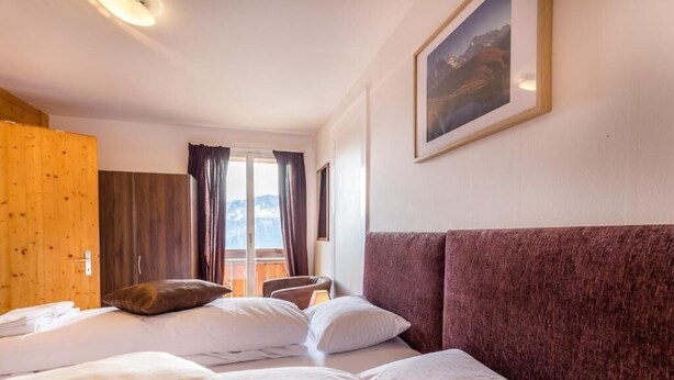 A hotel room with two beds, a wooden wardrobe, a large window with curtains, and a framed picture on the wall. - Hotel Sterne - Standard Apartment with Lake view (Beatenberg)