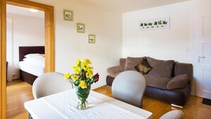 A living room with a brown sofa, a white table with a vase of flowers, and a bed in the background. - Hotel Sterne - Standard Apartment with Lake view (Beatenberg)