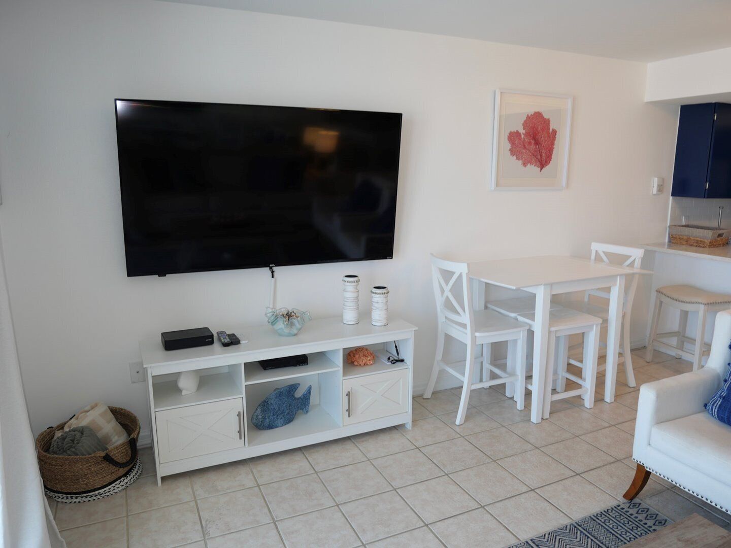 Una sala de estar con un televisor de pantalla plana montado en la pared, una mesa de comedor blanca con sillas, un sofá blanco y una pequeña cocina con armarios.