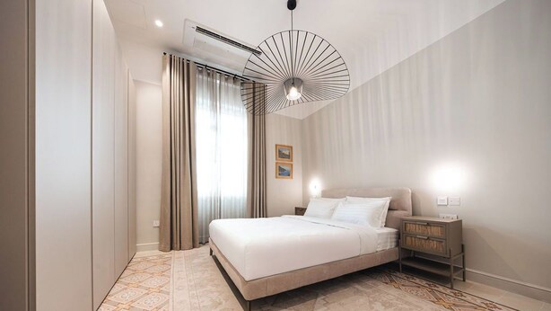 Un dormitorio con una cama, una mesita de noche, una lámpara de pared, una ventana con cortinas y un equipo de aire acondicionado.