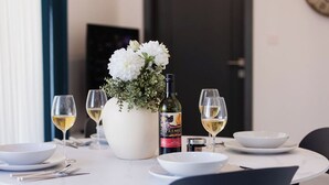 Une table à manger dressée avec de l’assiette blanche, des bols et des verres à vin, une bouteille de vin et un vase rempli de fleurs blanches.