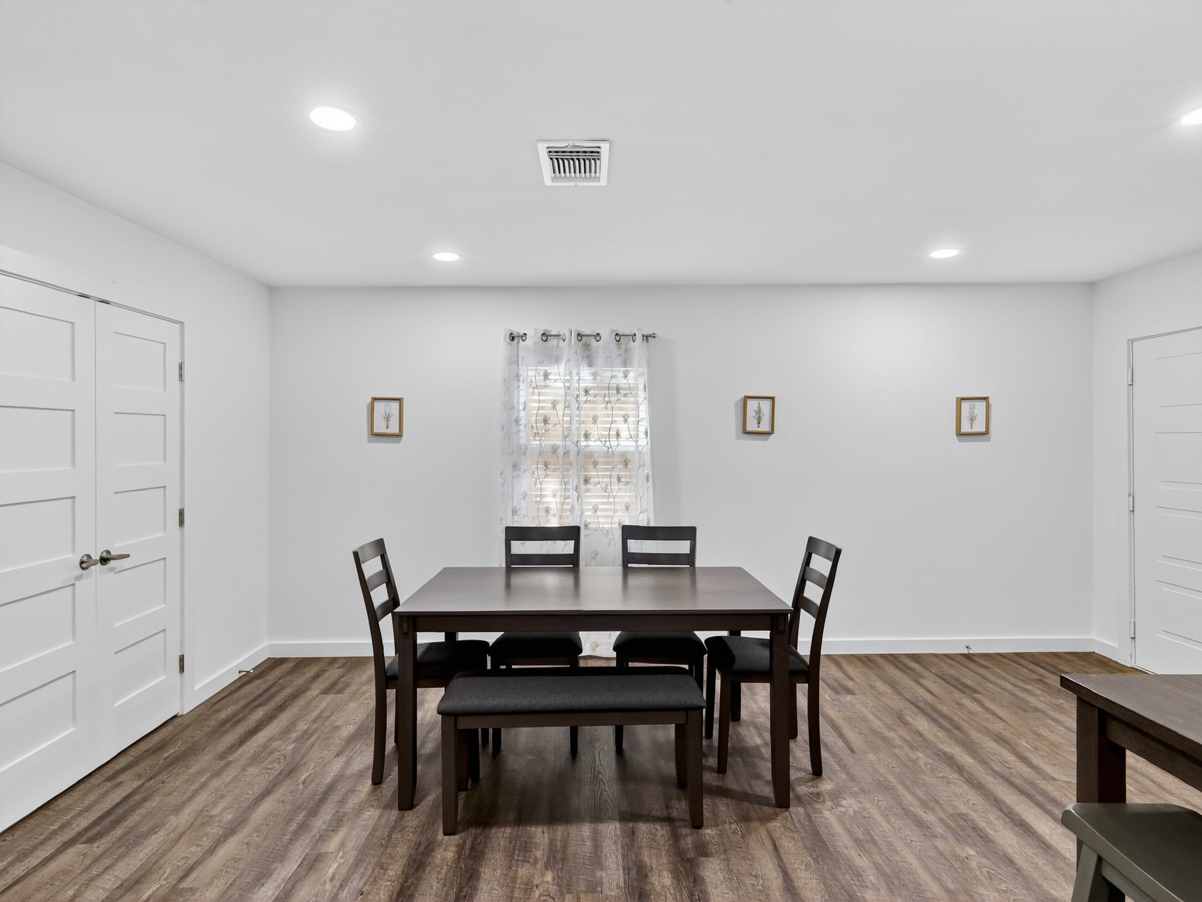 A dining room with a rectangular table, six chairs, a bench, and a window with a floral curtain.